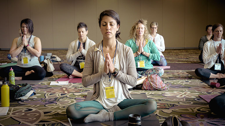 Photo: Group of Woman sitting cross-legged on carpeted floor with eyes closed and hands in prayer position, meditating.